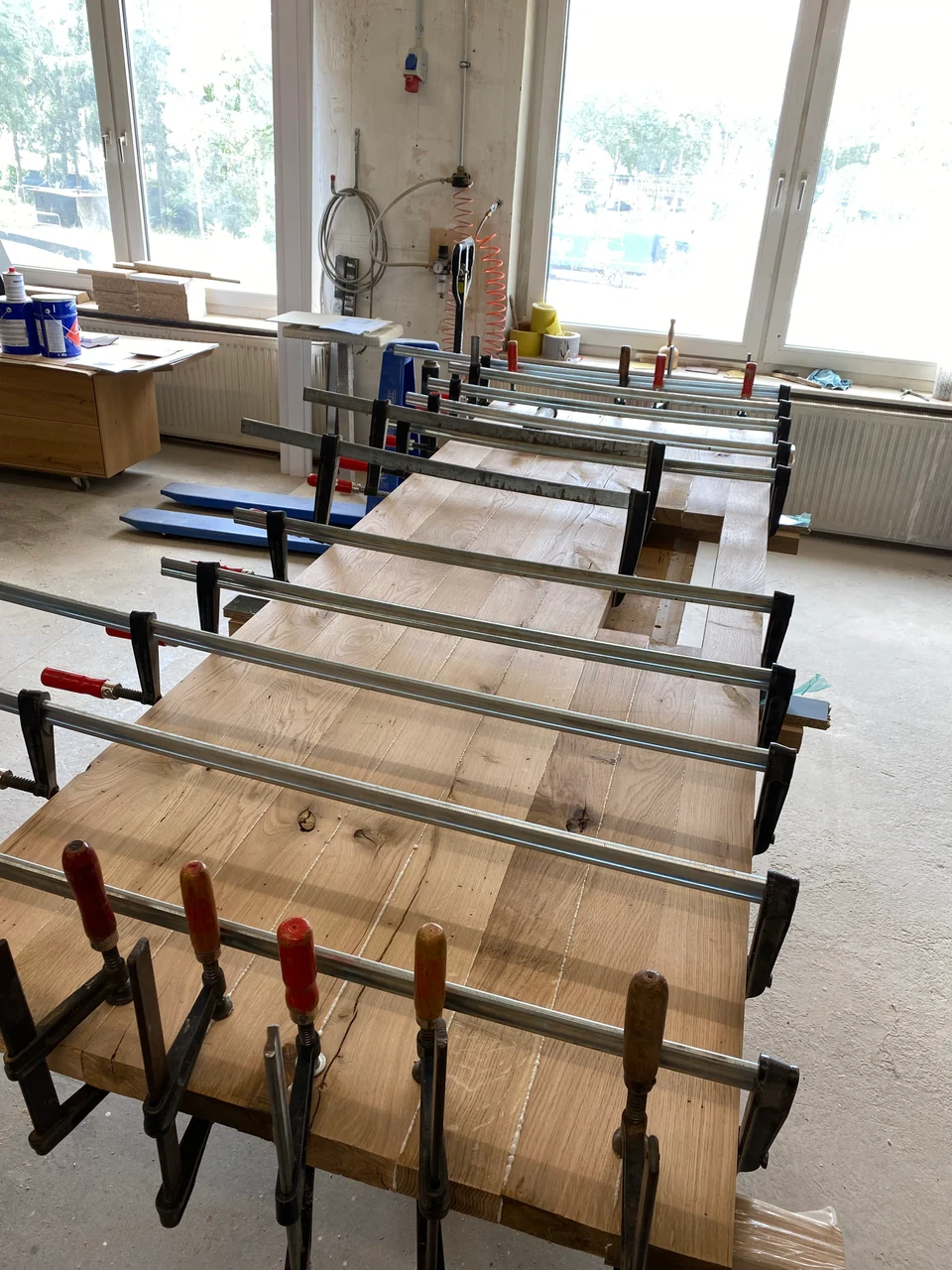 Werkstatt mit einer großen eingespannten Holzfläche, quer angesetzten Zwingen und Fenstern im Hintergrund